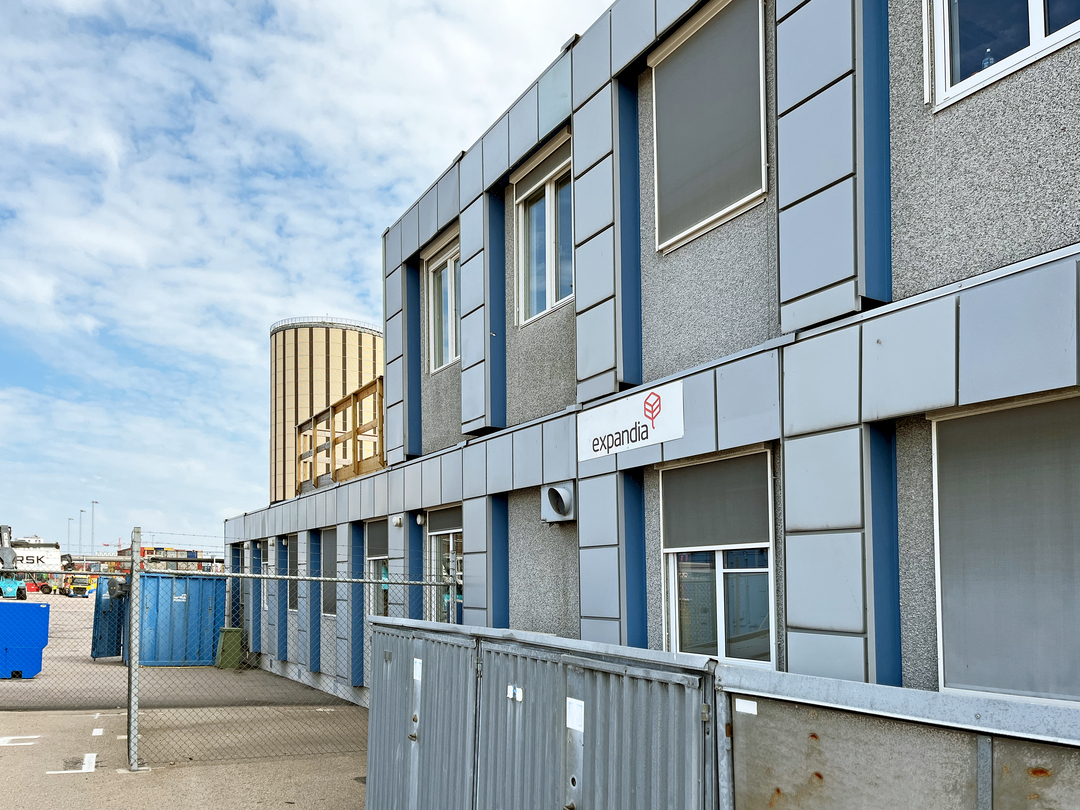 Helsingborgs hamn moduler i ett industriellt hamnområde, med grå fasadpaneler, fönster med neddragna solskydd och skylten ”expandia”, intill stängsel, containrar och en cylindrisk industribyggnad i bakgrunden under delvis molnig himmel.