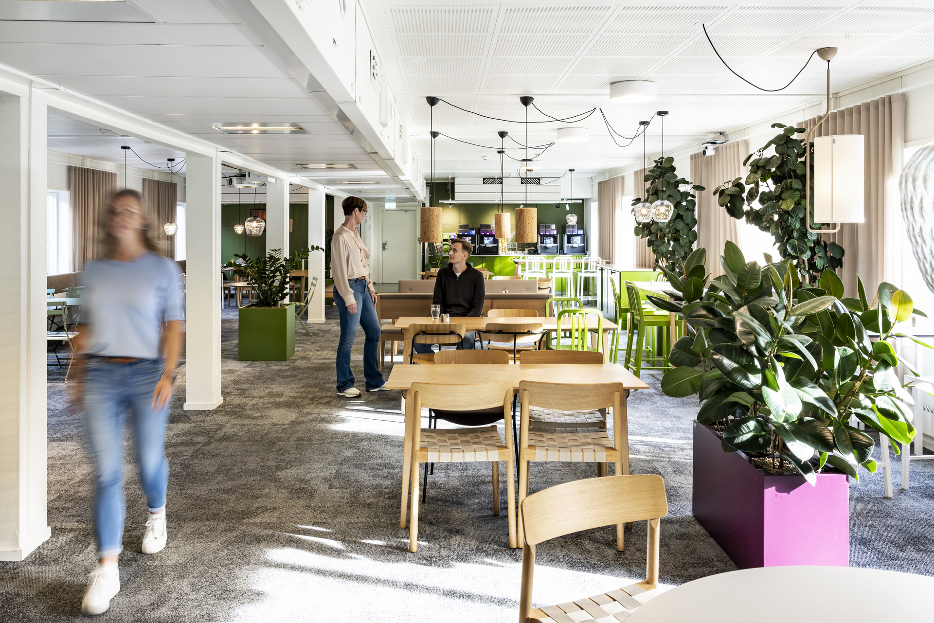 Öppen paus- och mötesyta i kontorsmoduler hos Polypeptide med träbord, gröna stolar, rikligt med växter och pendelbelysning, där medarbetare rör sig och samtalar i en ljus och modern arbetsmiljö. Kontorsmoduler från Expandia.