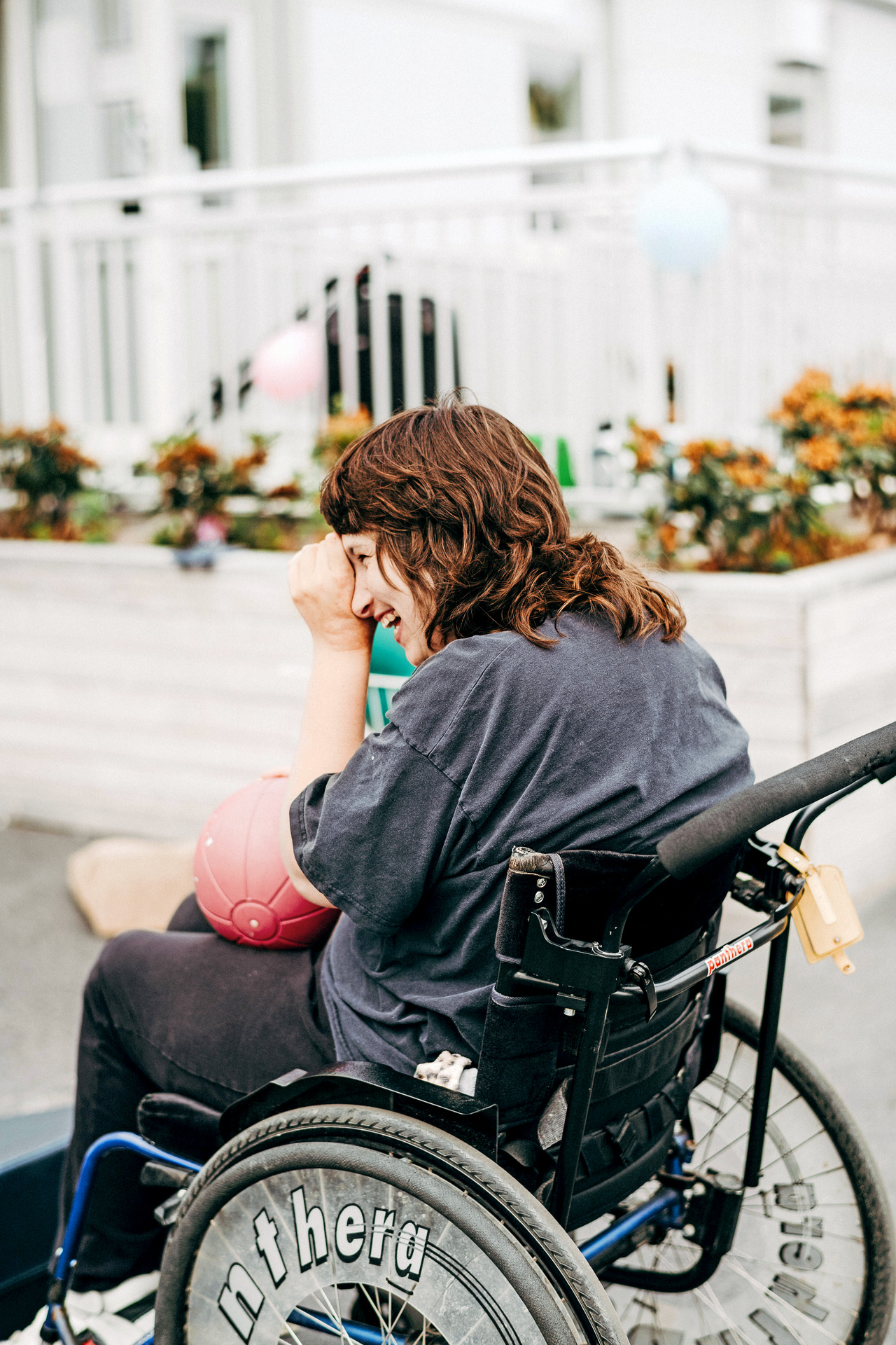 En person som sitter i rullstol skrattar och håller i en röd boll utomhus vid Insperia daglig LSS-verksamhet i Ekerö. I bakgrunden syns räcken, planteringar och en ljus, tillgänglig utemiljö.