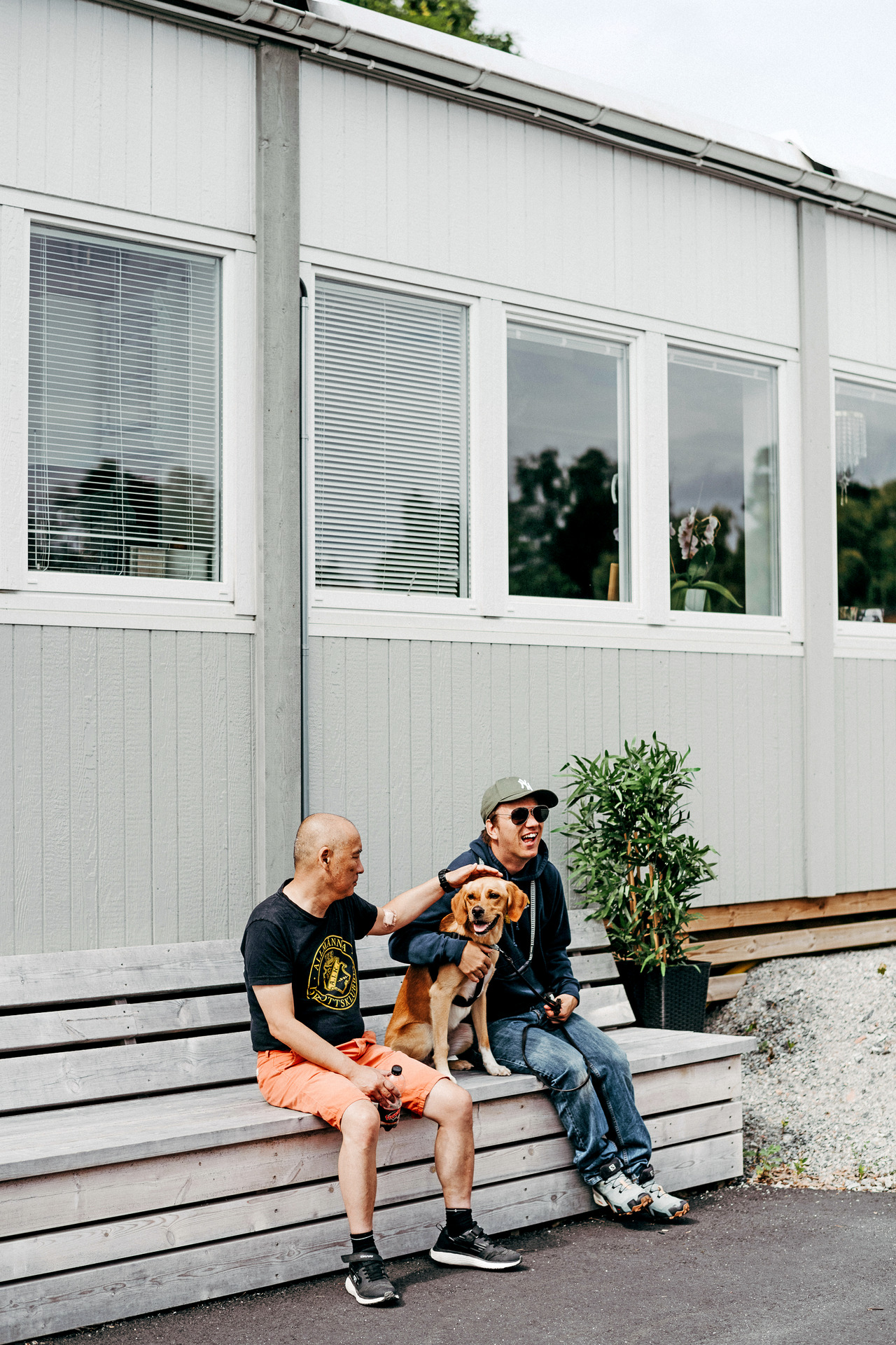 Två personer sitter på en bänk utomhus tillsammans med en hund. Den ena klappar hunden medan den andra skrattar och håller i kopplet.
