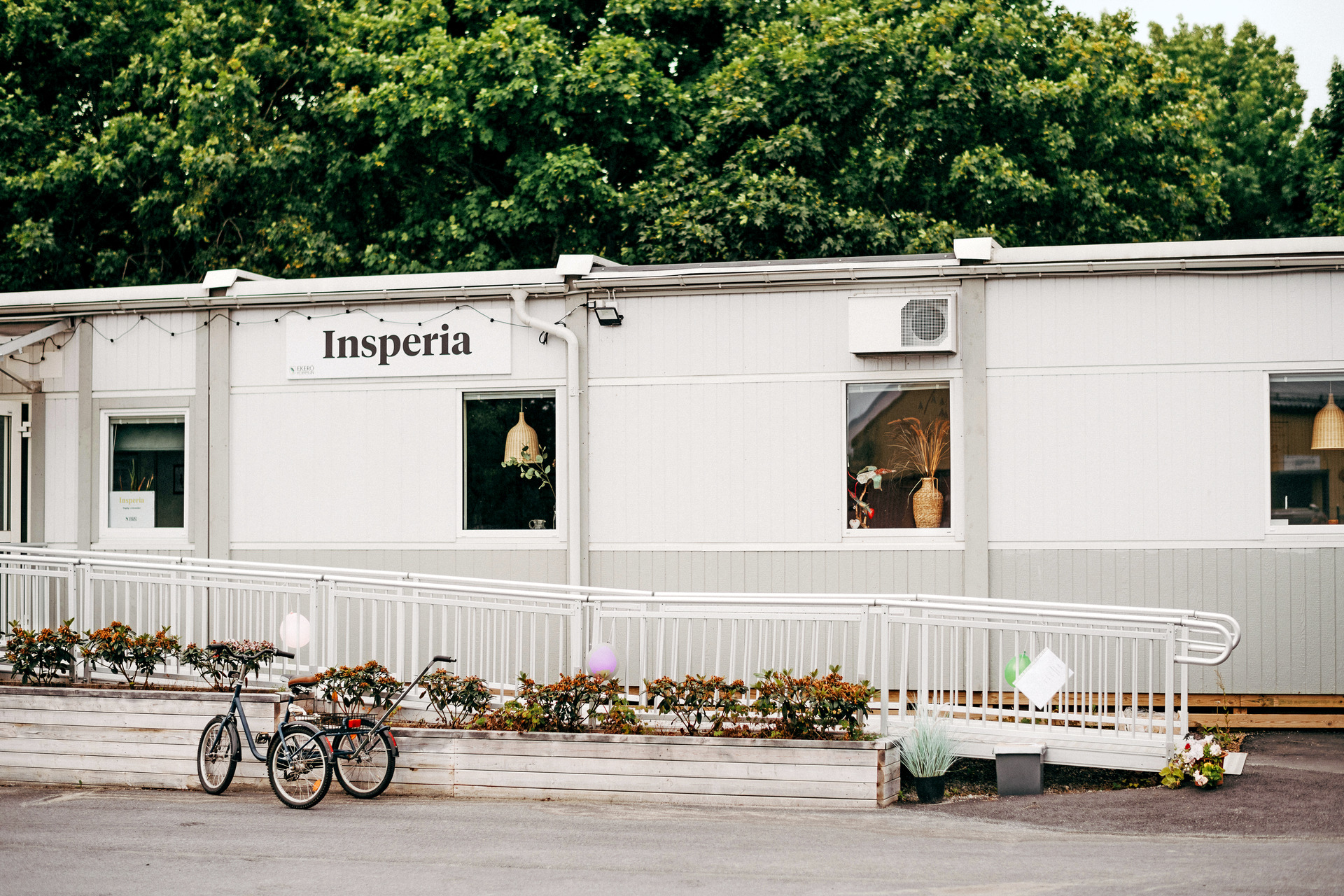 Exteriör av Insperia daglig LSS-verksamhet i Ekerö, inrymd i ljusa moduler. En tillgänglig ramp med räcke löper längs fasaden, med planteringar framför och grönskande träd i bakgrunden.
