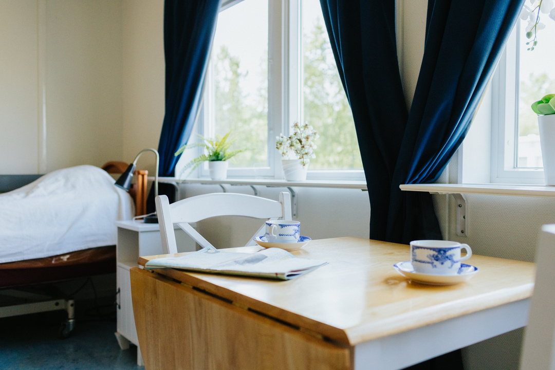Ett ljust rum vid Herrhagsgården vård- och omsorgsboende i Falun med säng, bord och två kaffekoppar. Rummet har fönster med mörka gardiner, växter på fönsterbrädan och möbler anpassade för vila och vardag.
