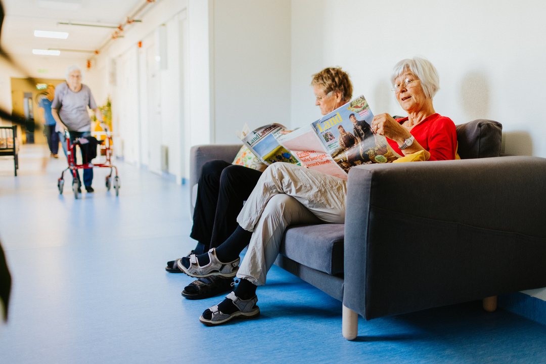 Två personer sitter i en soffa och läser tidningar i en korridor vid Herrhagsgården vård- och omsorgsboende i Falun. I bakgrunden syns en annan person som går med rollator i den ljusa inomhusmiljön.