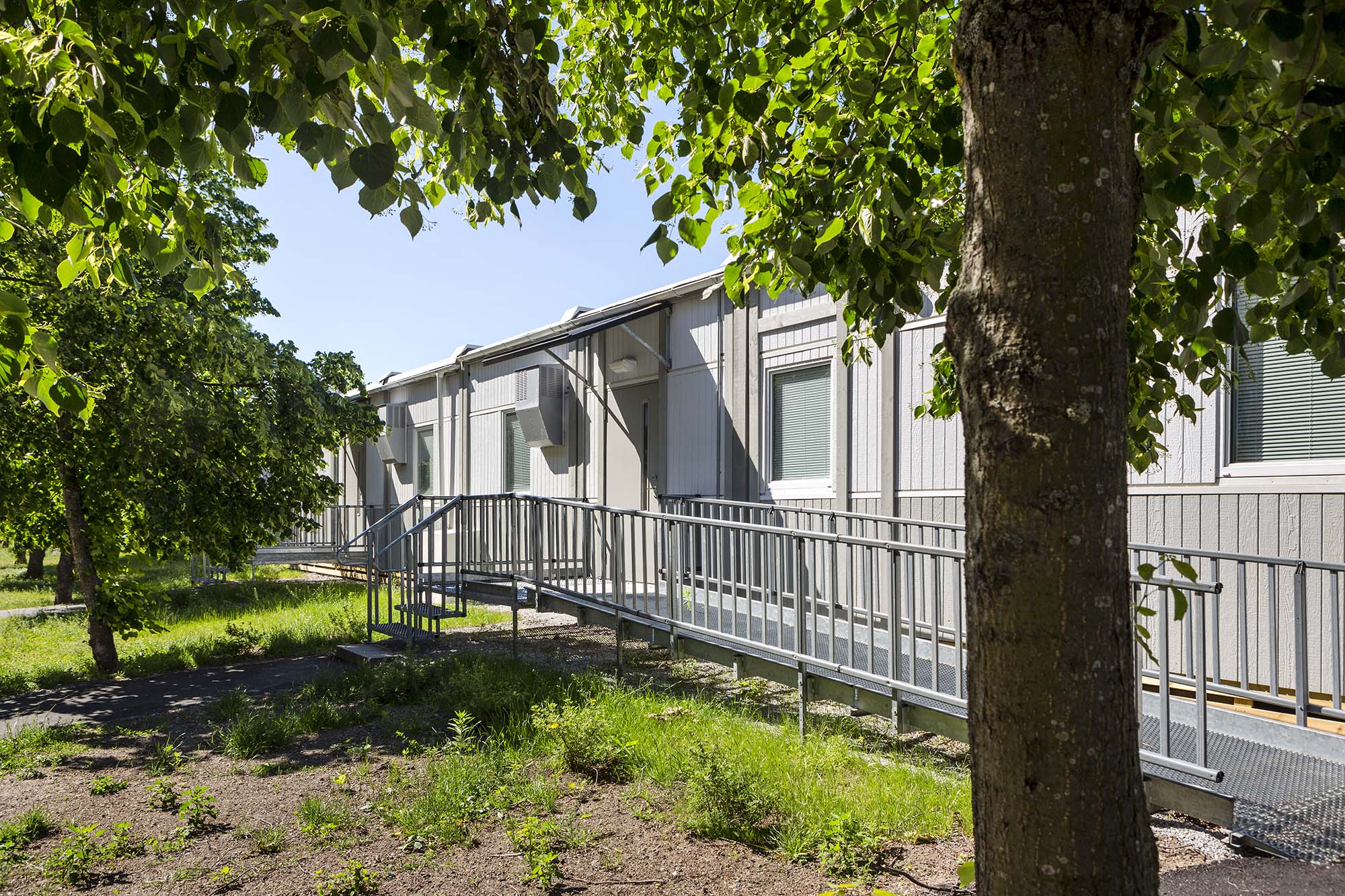 Exteriör av evakueringsboende i modulbyggnader med ljus fasad, lång ramp med räcke längs byggnaden och flera entréer. Byggnaden omges av grönska och träd i soligt väder.