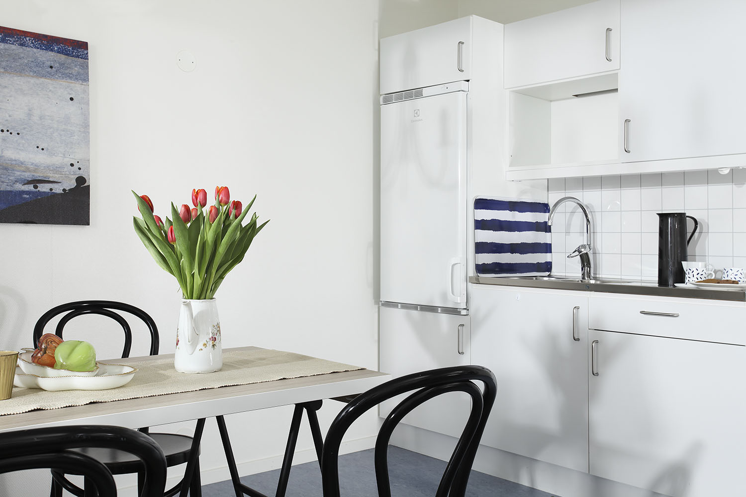 Pentry i boendemoduler från Expandia med vitt kök, kylskåp, diskbänk och matplats med svarta stolar samt tulpaner på bordet.